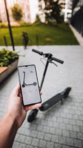 Close-up of a person's hand holding a smartphone with an app open displaying an icon of an electric scooter, with a real electric scooter in the background, suggesting the use of a ride-sharing app