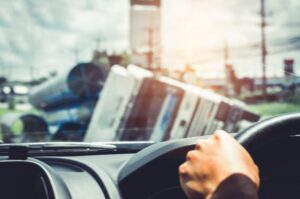 View from inside a car showing a driver's hand on the steering wheel, with a blurred vision of an overturned truck on the road ahead